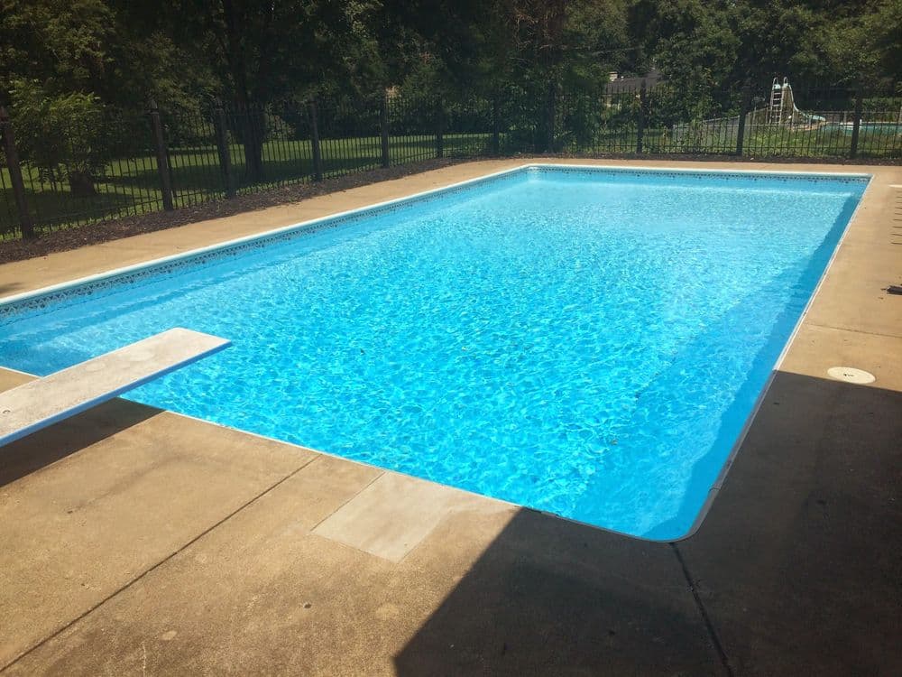 Bright blue swimming pool with diving board surrounded by a sunlit patio and lush greenery.